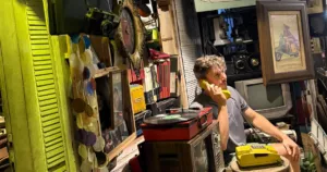 Jonas Thiry au téléphone avec un combiné jaune, entouré de téléviseurs, tourne-disque et objets rétro